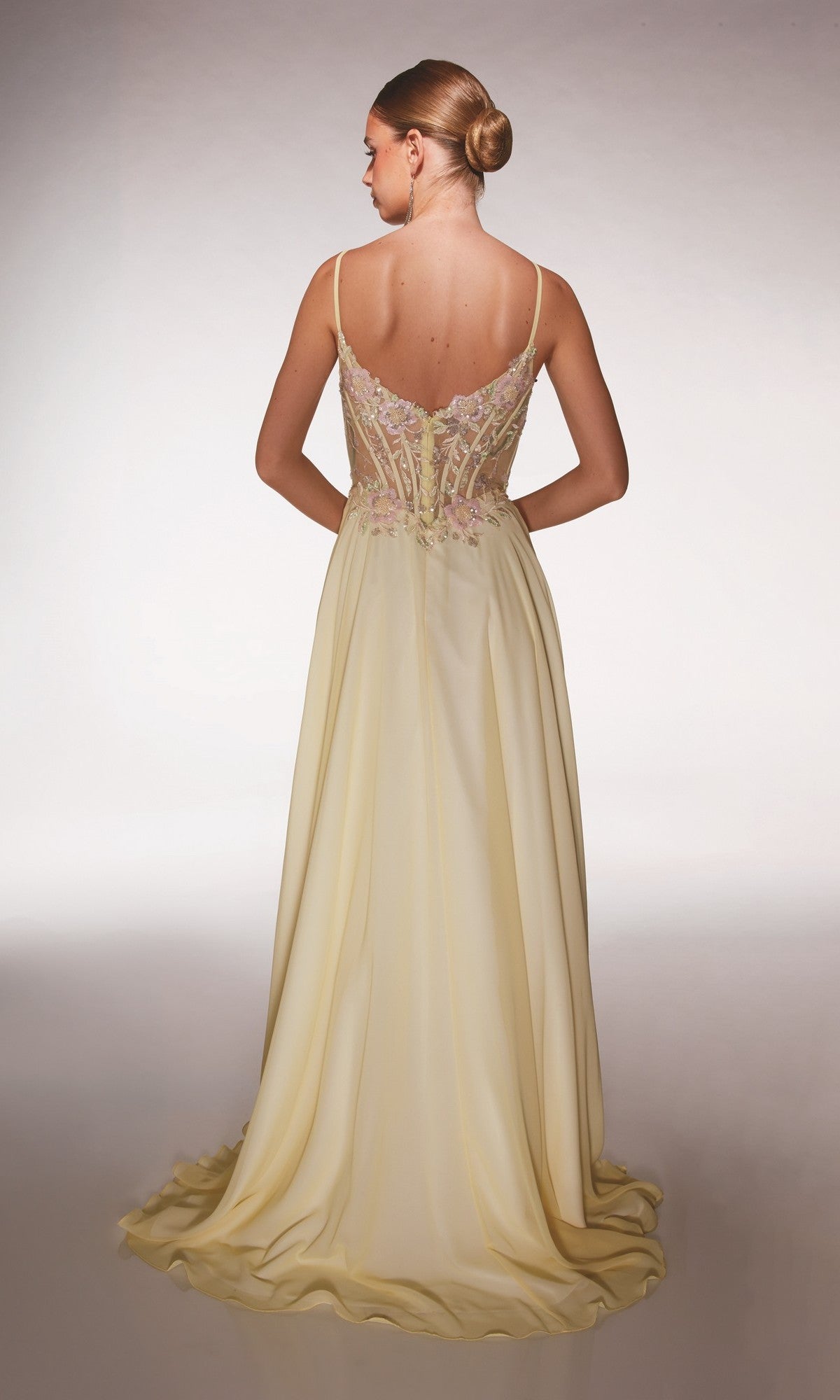 A woman stands with her back to the camera, wearing the Alyce 62198 long prom dress—a sleeveless, pale yellow gown with floral appliques on the bodice and a flowing chiffon A-line skirt.